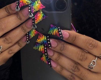 Puede incluir: Uñas acrílicas largas y coloridas con un diseño de estampado de cebra arcoíris. Las uñas están adornadas con bandas negras y pedrería. Algunas uñas tienen pedrería colgante. Las uñas están en una mano con un anillo de corazón plateado.
