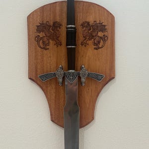 May include: A silver sword with a decorative handle and a wooden wall mount with two engraved griffins. The sword is mounted on the wall with the blade pointing downwards.