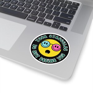 May include: A circular sticker with a yellow smiley face wearing blue and pink glasses. The sticker has the text "YOUR STRUGGLES DO NOT DEFINE YOU" in green, set against a black background. The sticker is on a laptop.