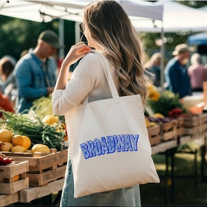 May include: A cream-colored tote bag with the word "BROADWAY" in blue, carried by a person at an outdoor market. The bag has a simple design, with the text prominently displayed. The market features wooden crates filled with produce.