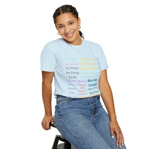 May include: A light blue t-shirt with a colorful design featuring song lyrics and phrases. The text includes "You're Beautiful", "Fully Dressed", "In Sleep", "He Sang", "To Me", "525,600 Minutes", "Raise A Star", "Anything can happen", "To Freedom", "Mama I'm a Big Girl Now".