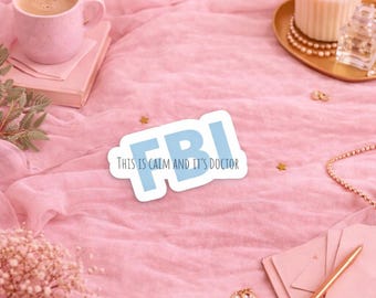 May include: A sticker with the letters "FBI" in light blue, with the text "THIS IS CALM AND IT'S DOCTOR" below it. The sticker is on a pink fabric surface, alongside a pink mug, a candle, and jewelry.