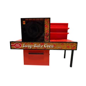 May include: A red and black Betty Crocker Easy-Bake Oven toy with a woven basket pattern and small red flower accents. The front panel features the text Betty Crocker Easy-Bake Oven by Kenner, a black oven door, and a red storage drawer below.