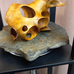 Puede incluir: Una escultura de madera con un dise&ntilde;o abstracto &uacute;nico. La escultura est&aacute; hecha de madera marr&oacute;n claro y tiene un acabado liso y pulido. Se muestra sobre una base de piedra gris oscuro.