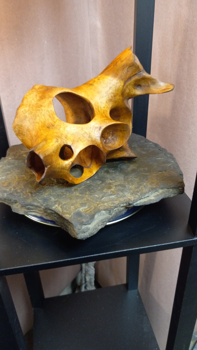Puede incluir: Una escultura de madera con un dise&ntilde;o abstracto &uacute;nico. La escultura est&aacute; hecha de madera marr&oacute;n claro y tiene un acabado liso y pulido. Se muestra sobre una base de piedra gris oscuro.