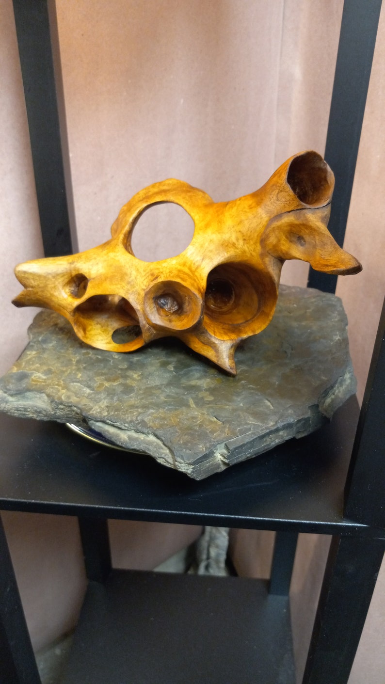 Puede incluir: Una escultura de madera de una concha marina con detalles intrincados y un acabado de madera natural. La escultura se exhibe sobre una base de pizarra gris oscuro.
