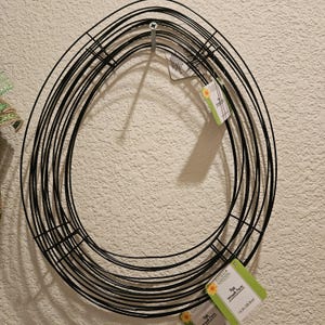May include: Black metal egg-shaped wreath form hanging on a wall. The wreath form is made of multiple layers of wire. The product tag reads "Egg Wreath Form" and measures 15.3 in (38.8 cm).
