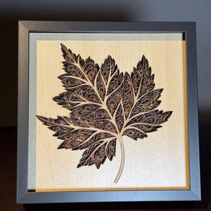 Puede incluir: Una talla de madera enmarcada de una hoja de arce detallada con intrincados patrones en tonos marrones y rojos.