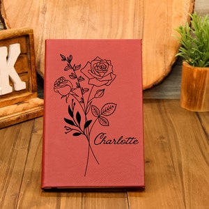 Puede incluir: Un diario burdeos con un dise&ntilde;o floral de rosas y el nombre "Charlotte" grabado en la portada. El diario est&aacute; cerrado y se encuentra de pie sobre una superficie de madera. El dise&ntilde;o presenta dos rosas y hojas.