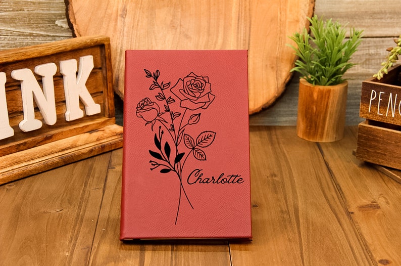 Puede incluir: Un diario burdeos con un dise&ntilde;o floral de rosas y el nombre "Charlotte" grabado en la portada. El diario est&aacute; cerrado y se encuentra de pie sobre una superficie de madera. El dise&ntilde;o presenta dos rosas y hojas.