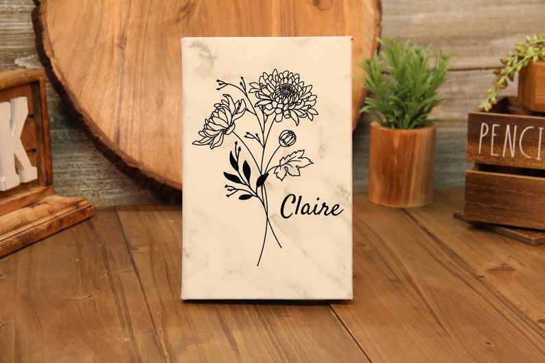 Puede incluir: Un lienzo rectangular con un patr&oacute;n similar al m&aacute;rmol presenta un dise&ntilde;o floral negro y el nombre "Claire". La obra de arte incluye dibujos detallados de flores, hojas y tallos. El lienzo se exhibe sobre una superficie de madera.