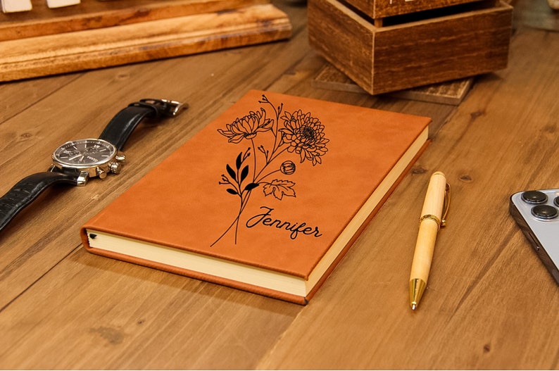 Puede incluir: Un diario de cuero marr&oacute;n con un dise&ntilde;o floral y el nombre "Jennifer" grabado en la portada. Un reloj de cuero negro, un bol&iacute;grafo de madera y un tel&eacute;fono inteligente tambi&eacute;n est&aacute;n sobre la superficie de madera.