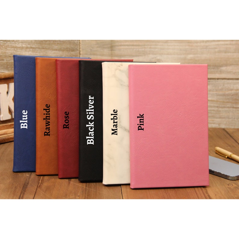 Puede incluir: Una colecci&oacute;n de diarios encuadernados en cuero en varios colores, incluyendo azul, cuero crudo, rosa, negro, m&aacute;rmol y rosa. Cada diario tiene una cubierta texturizada y el nombre del color impreso verticalmente. Un bol&iacute;grafo y un tel&eacute;fono est&aacute;n sobre la superficie de madera.