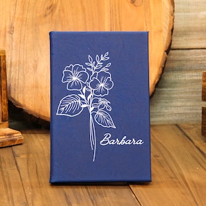 Puede incluir: Un diario azul marino con un dise&ntilde;o floral blanco y el nombre "Barbara" en cursiva. El diario rectangular parece ser de cuero. El fondo incluye elementos de madera.