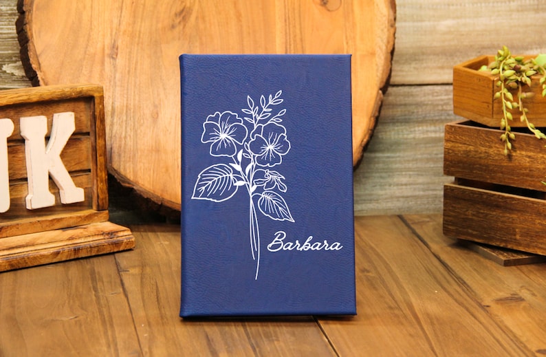 Puede incluir: Un diario azul marino con un dise&ntilde;o floral blanco y el nombre "Barbara" en cursiva. El diario rectangular parece ser de cuero. El fondo incluye elementos de madera.