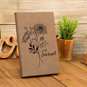Puede incluir: Un diario de color beige con un dise&ntilde;o floral y el nombre "Sarah" grabado en la portada. El diario est&aacute; de pie sobre una superficie de madera, con una peque&ntilde;a planta y adornos de madera en el fondo.