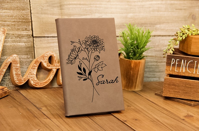 Puede incluir: Un diario de color beige con un dise&ntilde;o floral y el nombre "Sarah" grabado en la portada. El diario est&aacute; de pie sobre una superficie de madera, con una peque&ntilde;a planta y adornos de madera en el fondo.