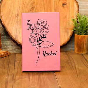 Puede incluir: Un diario rosa con un dise&ntilde;o floral negro y el nombre "Rachel" grabado. El diario est&aacute; de pie sobre una superficie de madera. El dise&ntilde;o floral incluye una flor grande, capullos y hojas.