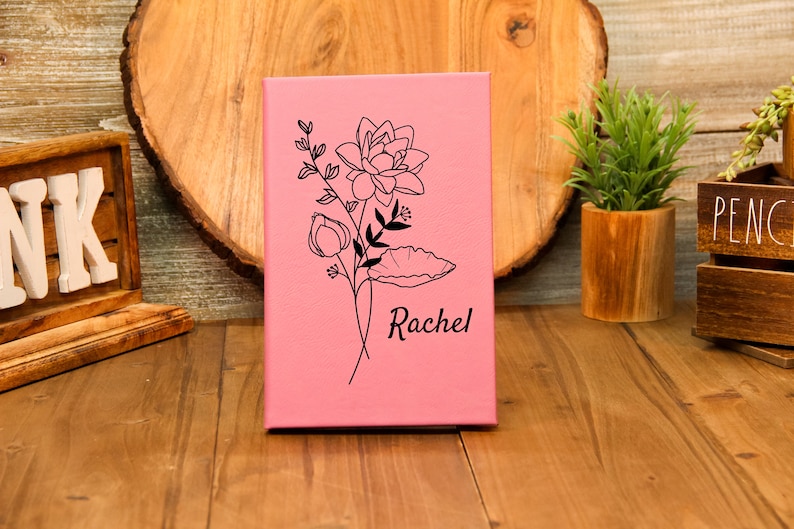 Puede incluir: Un diario rosa con un dise&ntilde;o floral negro y el nombre "Rachel" grabado. El diario est&aacute; de pie sobre una superficie de madera. El dise&ntilde;o floral incluye una flor grande, capullos y hojas.