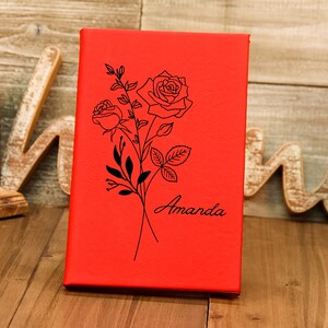 Puede incluir: Un diario rojo con un dise&ntilde;o floral de rosas negras y el nombre "Amanda" en escritura cursiva. La cubierta tiene una textura lisa. El diario est&aacute; sobre una superficie de madera.