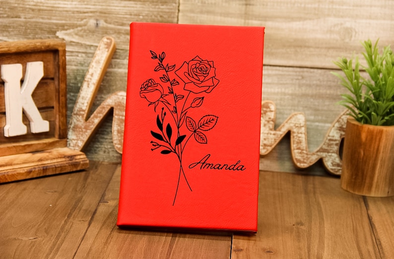 Puede incluir: Un diario rojo con un dise&ntilde;o floral de rosas negras y el nombre "Amanda" en escritura cursiva. La cubierta tiene una textura lisa. El diario est&aacute; sobre una superficie de madera.