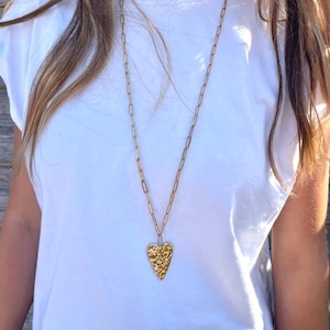 Peut inclure: Un long collier doré avec un pendentif en forme de cœur texturé. Le pendentif a un aspect martelé. Le collier est porté sur un t-shirt blanc.