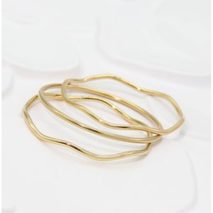 Puede incluir: Un conjunto de tres anillos de metal en tono dorado, cada uno con un diseño de banda circular suave, continuo y ondulado. Los anillos están apilados superponiéndose ligeramente, mostrando una superficie pulida y reflectante.