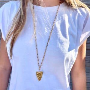 Peut inclure: Un long collier doré avec un pendentif en forme de cœur texturé. Le pendentif a une surface rugueuse et inégale. Le collier est porté sur un t-shirt blanc.