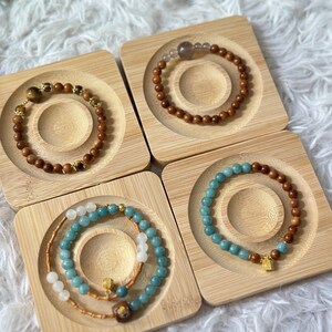 May include: Four wooden bracelets with different coloured beads and gold accents displayed on small wooden squares. The bracelets are made of brown wood, blue beads, and white beads.