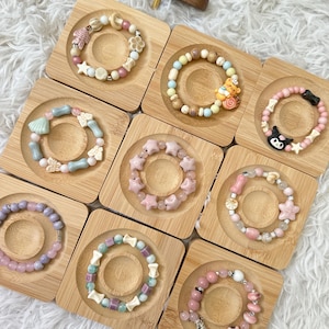 May include: Ten colorful beaded bracelets displayed on small wooden stands. The bracelets feature various charms, including stars, seashells, butterflies, and cartoon characters. The bracelets are made with pink, blue, green, and white beads.