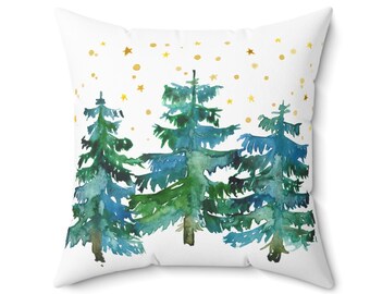 Pode incluir: Almofada quadrada branca com um desenho em aquarela de três árvores perenes em azul e verde. Estrelas douradas estão espalhadas no fundo branco, criando um tema festivo e invernal.