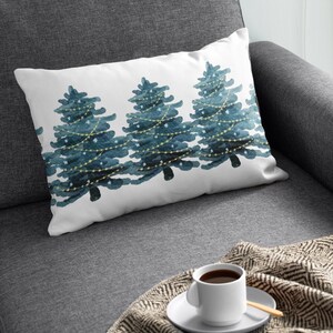 May include: A white throw pillow with a watercolor design of three blue Christmas trees with white lights and stars. The pillow is on a gray couch with a blanket and a white coffee cup.