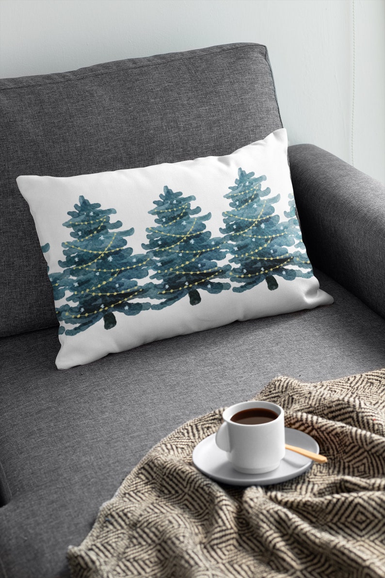 May include: A white throw pillow with a watercolor design of three blue Christmas trees with white lights and stars. The pillow is on a gray couch with a blanket and a white coffee cup.