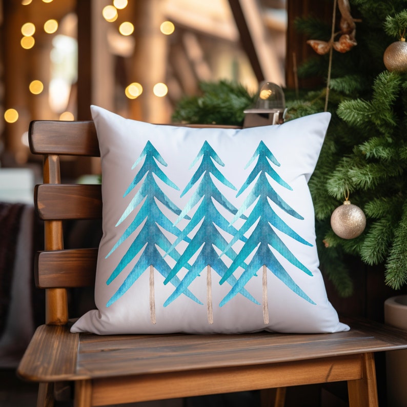 Pode incluir: Almofada quadrada branca com um desenho em aquarela de tr&ecirc;s &aacute;rvores de Natal azul-esverdeadas estilizadas. As &aacute;rvores t&ecirc;m troncos castanhos e est&atilde;o dispostas em fila. A almofada est&aacute; numa cadeira de madeira, com uma &aacute;rvore de Natal ao fundo.