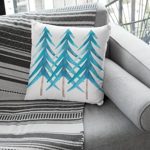 Pode incluir: Almofada quadrada branca com um desenho em aquarela de tr&ecirc;s pinheiros azul-esverdeados sobrepostos com troncos marrons. A almofada est&aacute; colocada em um sof&aacute; cinza com uma manta listrada em preto e branco.