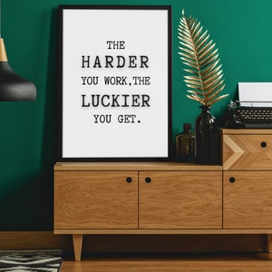 Op de afbeelding: Een ingelijste print met de tekst "THE HARDER YOU WORK, THE LUCKIER YOU GET." staat boven een houten kast. Een zwarte hanglamp hangt aan het plafond. Een gouden blad en een vintage typemachine staan op de kast.
