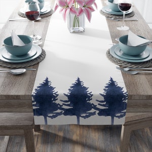 May include: White table runner with a navy blue watercolor design of three pine trees. The runner is on a wooden table with place settings and a vase of pink flowers.