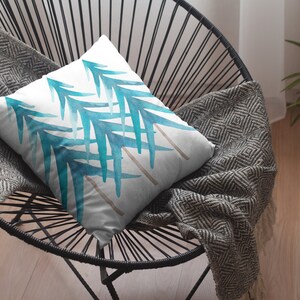 Pode incluir: Uma almofada quadrada com fundo branco e um desenho em aquarela de folhas azul-esverdeadas. A almofada est&aacute; em uma cadeira preta com um cobertor cinza estampado.