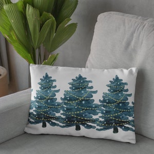 May include: A white throw pillow with a watercolor design of three blue Christmas trees with yellow lights. The pillow is on a gray couch.