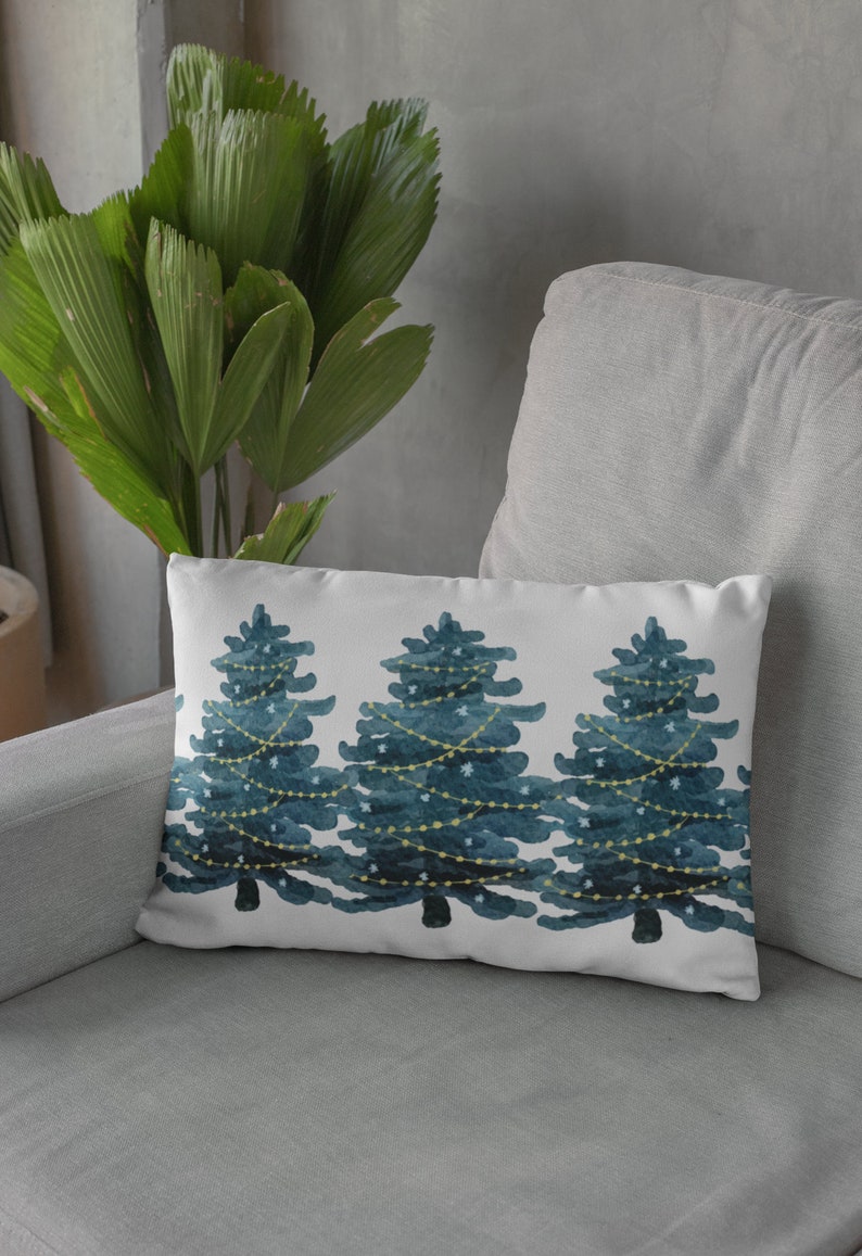 May include: A white throw pillow with a watercolor design of three blue Christmas trees with yellow lights. The pillow is on a gray couch.