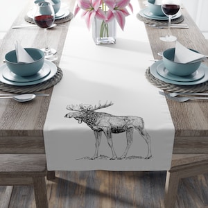 May include: White table runner featuring a detailed black and white illustration of a moose. The runner is placed on a wooden table set for a meal, with plates, bowls, silverware, and wine glasses. A vase of pink flowers is in the center.