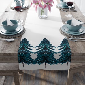 May include: White table runner with a teal blue watercolor pine tree design. The runner is on a wooden table with place settings.