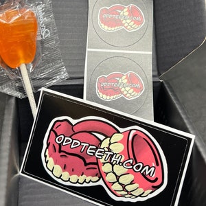 May include: An open black box containing an orange lollipop in a clear wrapper, stickers with a cartoon teeth design, and a rectangular sticker with the text "ODDTEETH.COM" and a cartoon teeth design.