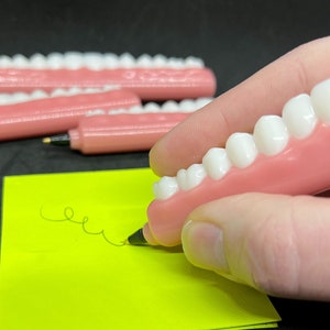 May include: A novelty pen shaped like a set of teeth, with a pink base and white teeth. The pen is writing on a neon yellow sticky note. The text "Round - Opaque" is printed on the sticky note.