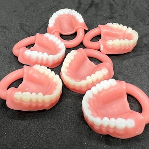 May include: A collection of novelty teeth-shaped items. Each item features a pink gum base with a set of white teeth and a pink handle. The items are arranged on a dark surface.