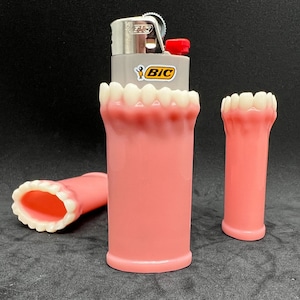 May include: A gray BIC lighter with a red ignition button is encased in a pink, tooth-shaped holder. Two additional pink holders with white teeth are also present, one upright and one lying down. The holders are designed to resemble a set of teeth.