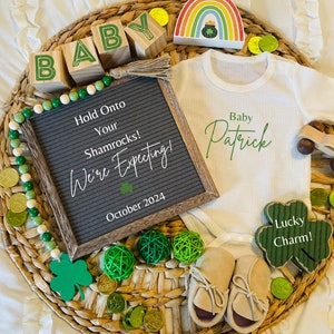 May include: A baby announcement with a white onesie that says "Baby Patrick" in green script. The onesie is surrounded by green shamrocks, gold coins, and wooden blocks that spell out "BABY". A letter board says "Hold Onto Your Shamrocks! We're Expecting! October 2024".