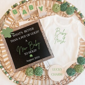 May include: A white onesie with the text "Baby Irish" in green script. A wooden sign with the text "What's better than a pot of gold? A New Baby to hold! August 2024" and a wooden circle with the text "Lucky Charm". The items are arranged on a woven wicker tray with green and white beads around the edge.