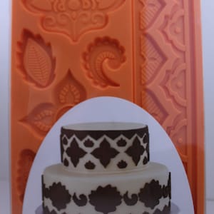 May include: Orange silicone mold with various intricate designs, including a large floral pattern, paisley shapes, and a border design. A two-tiered cake decorated with the designs from the mold is shown.