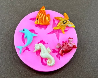May include: Pink silicone mold with five cavities shaped like a seahorse, a dolphin, a starfish, a crab, and a fish. The mold is filled with colorful resin casts of each shape.